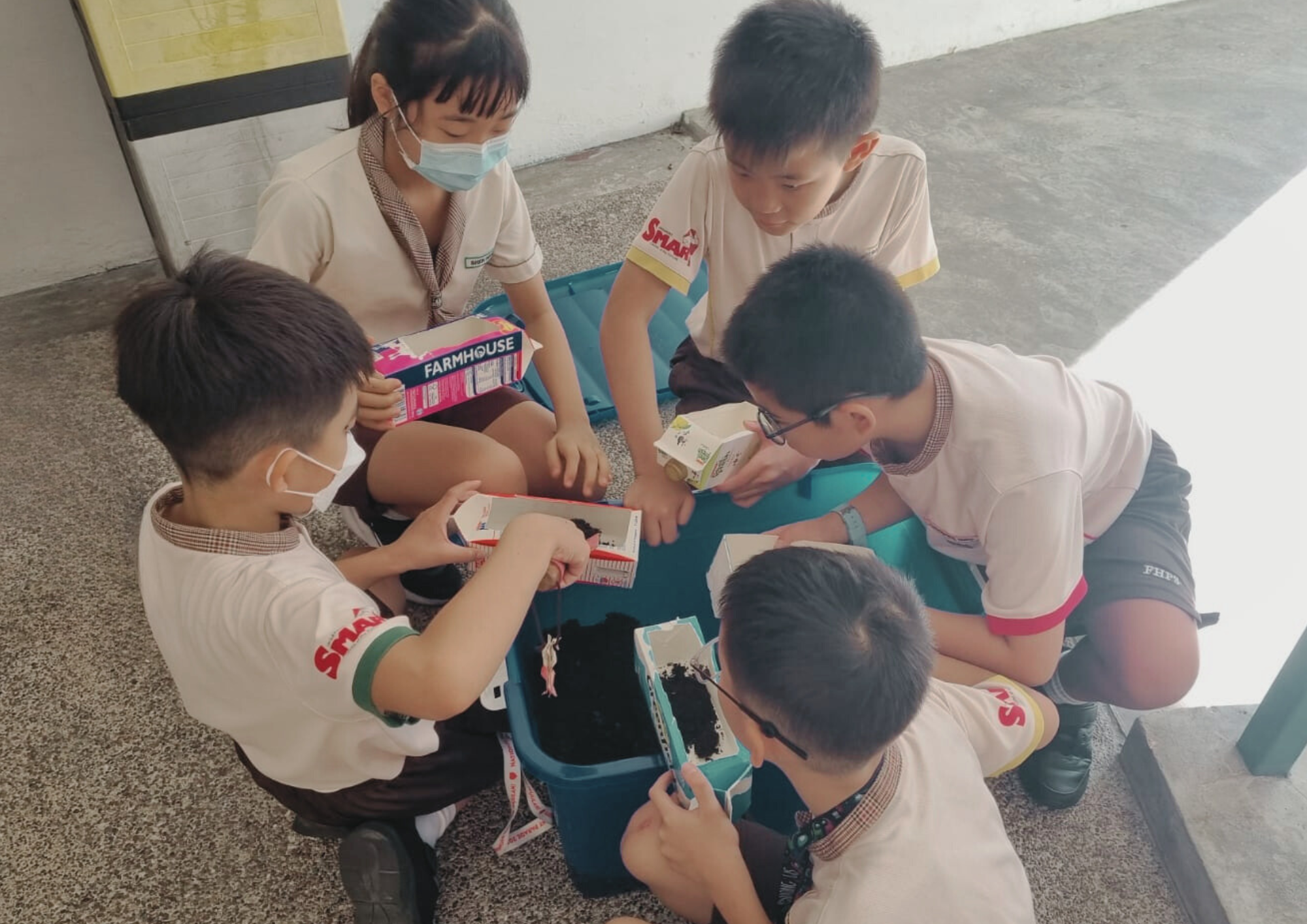 Five school children in uniforms sit around a teal bin, filling milk cartons with dark soil outdoors.